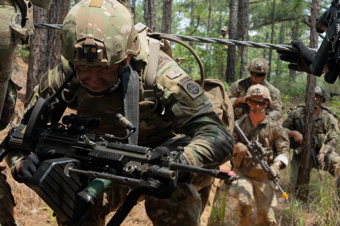 A soldier in tactical gear holds a weapon and ducks under wire in a forest setting while other soldiers follow.