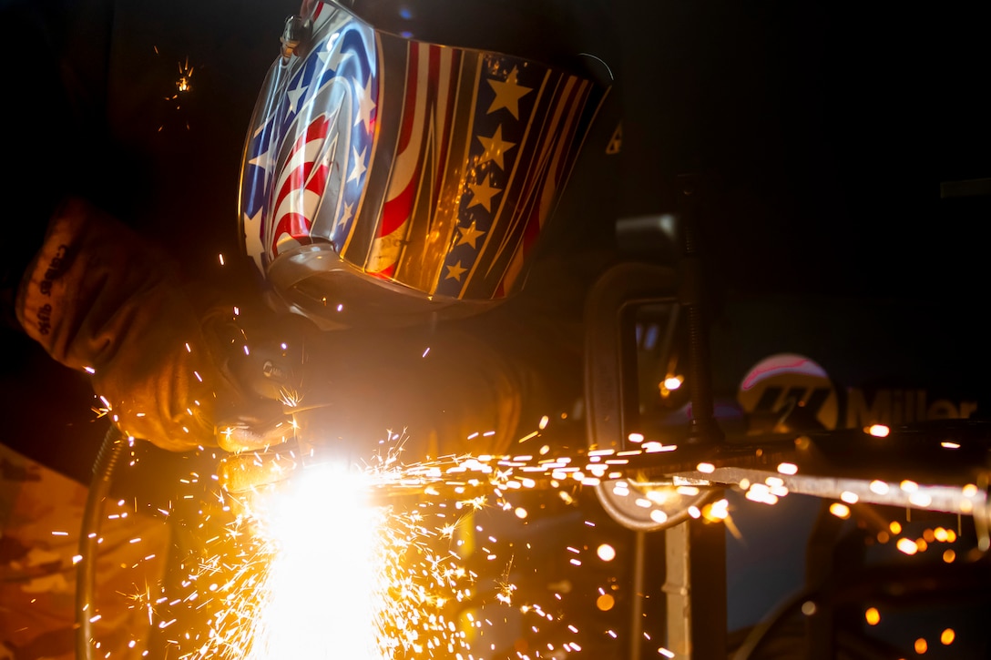 An airman with a welding helmet and gloves uses a plasma-cutting tool, causing sparks to fly above metal equipment.