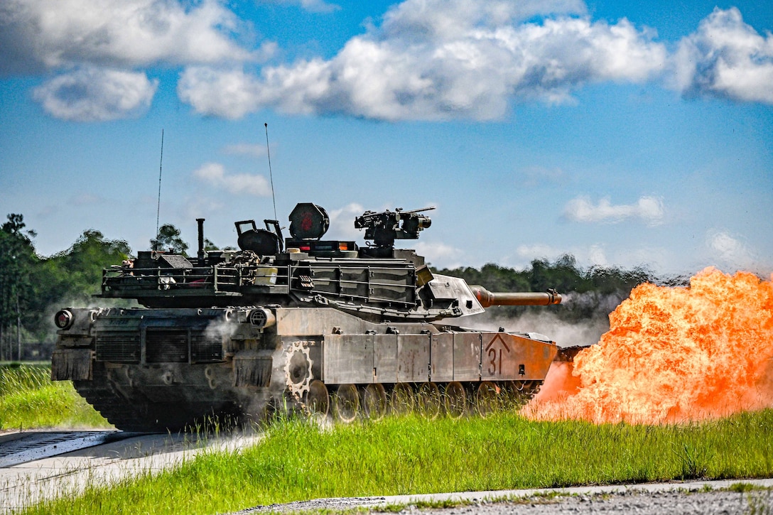 A tank is shown next to a blaze of fire in a field under blue, partly cloudy skies.