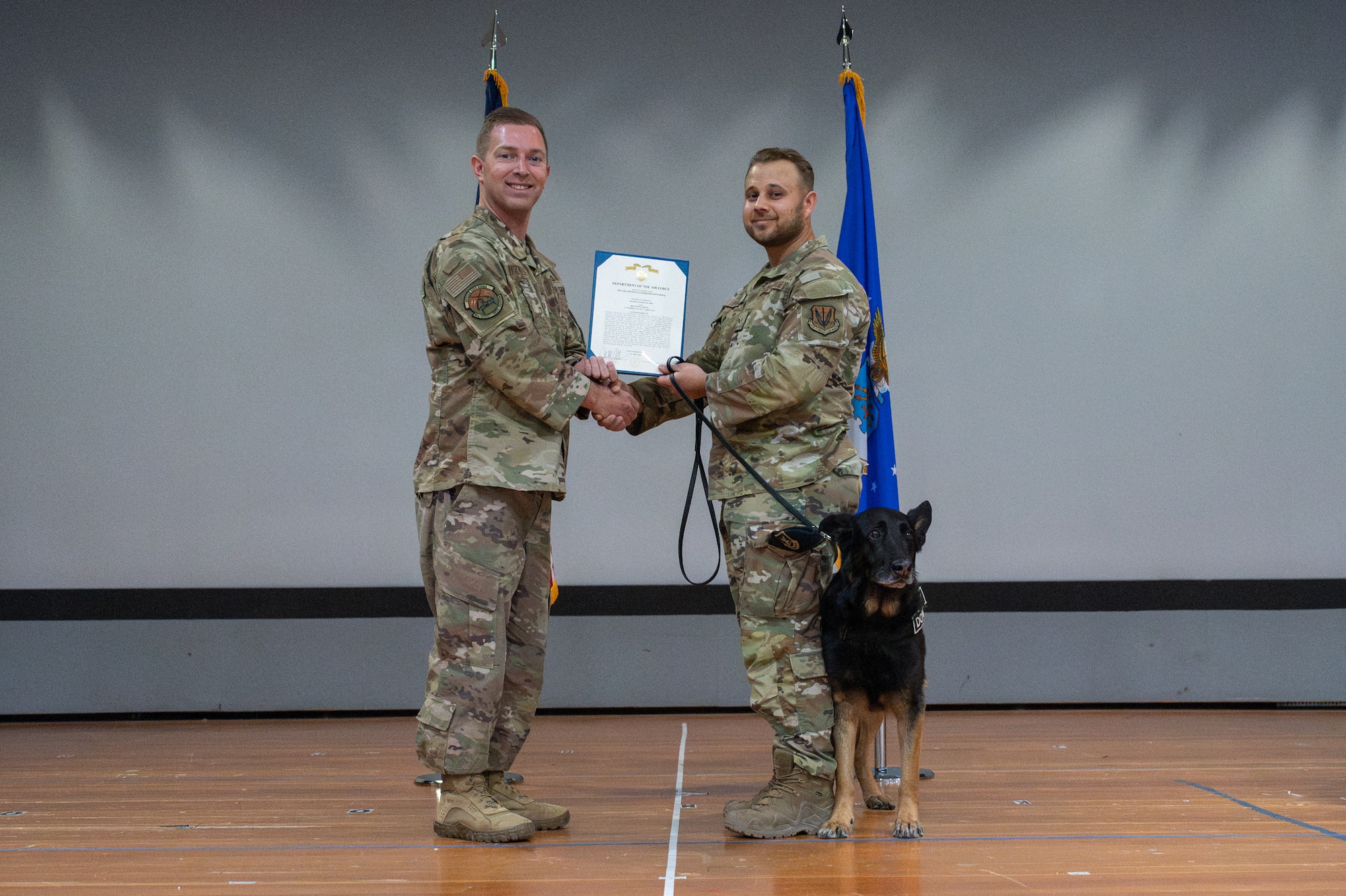 Two airman hold an award on a stage with a dog next to them.