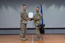 Two airman hold an award on a stage with a dog next to them.