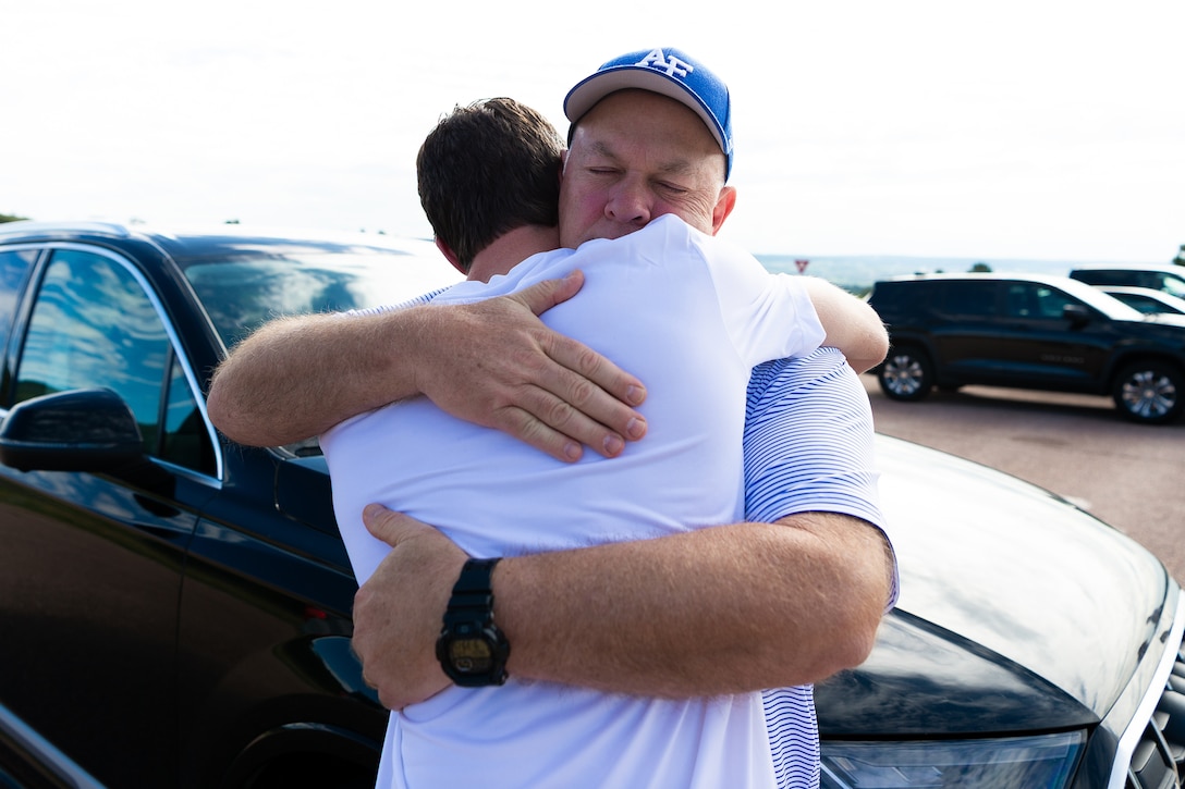A person hugs another person outside in front of parked cars.