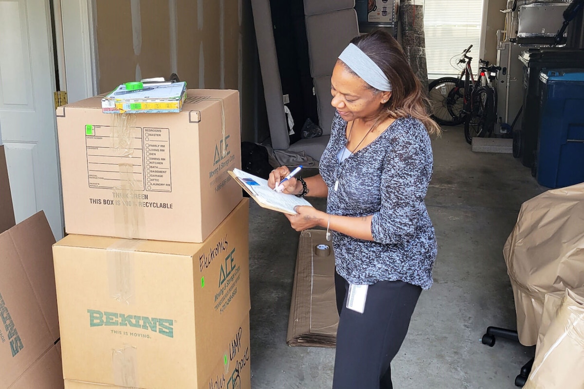 A woman with a clipboard stands next to three large cardboard boxes and checks items on a list.