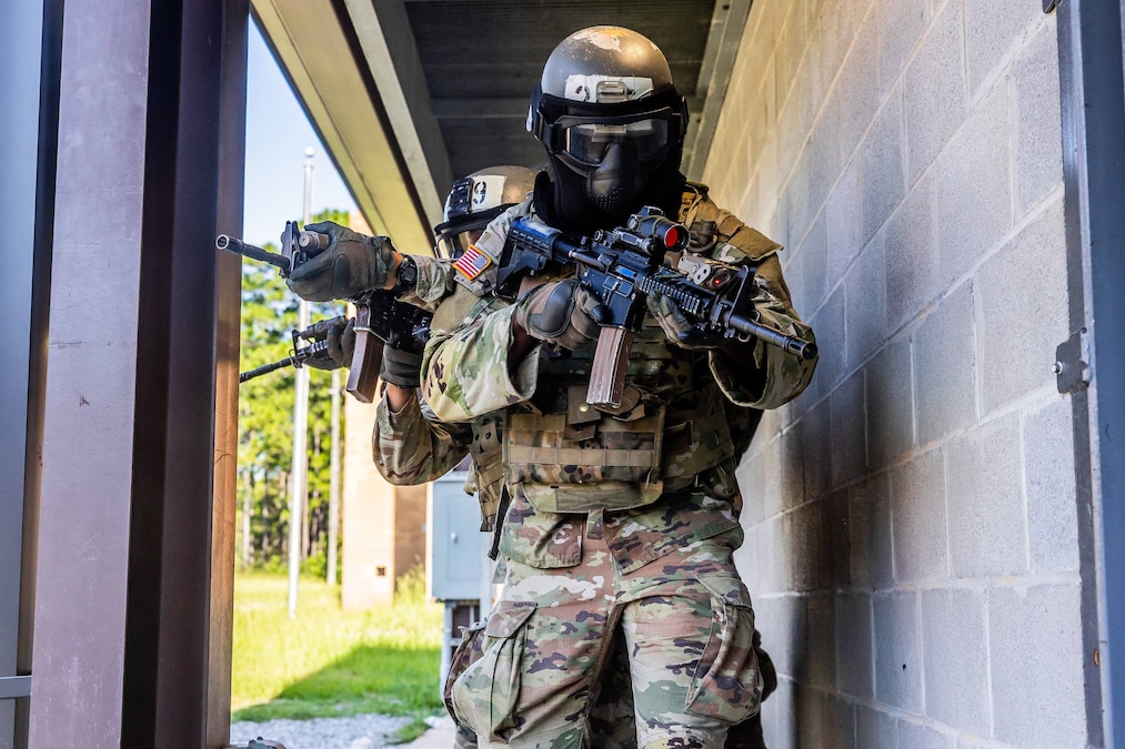 Two soldiers in tactical gear and helmets aim weapons in an open doorway next to an external wall.