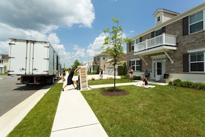 A man rolls a large item into a house while three people sit on the lawn and watch.