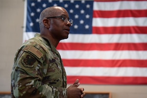A man in military uniform stands in front of a U.S. flag.