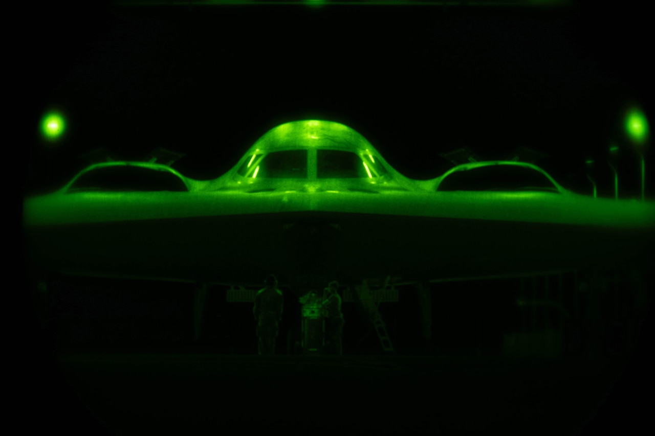 The center section of an aircraft is visible with green illumination almost entirely through the use of night-vision goggles. There are two airmen working underneath the aircraft.