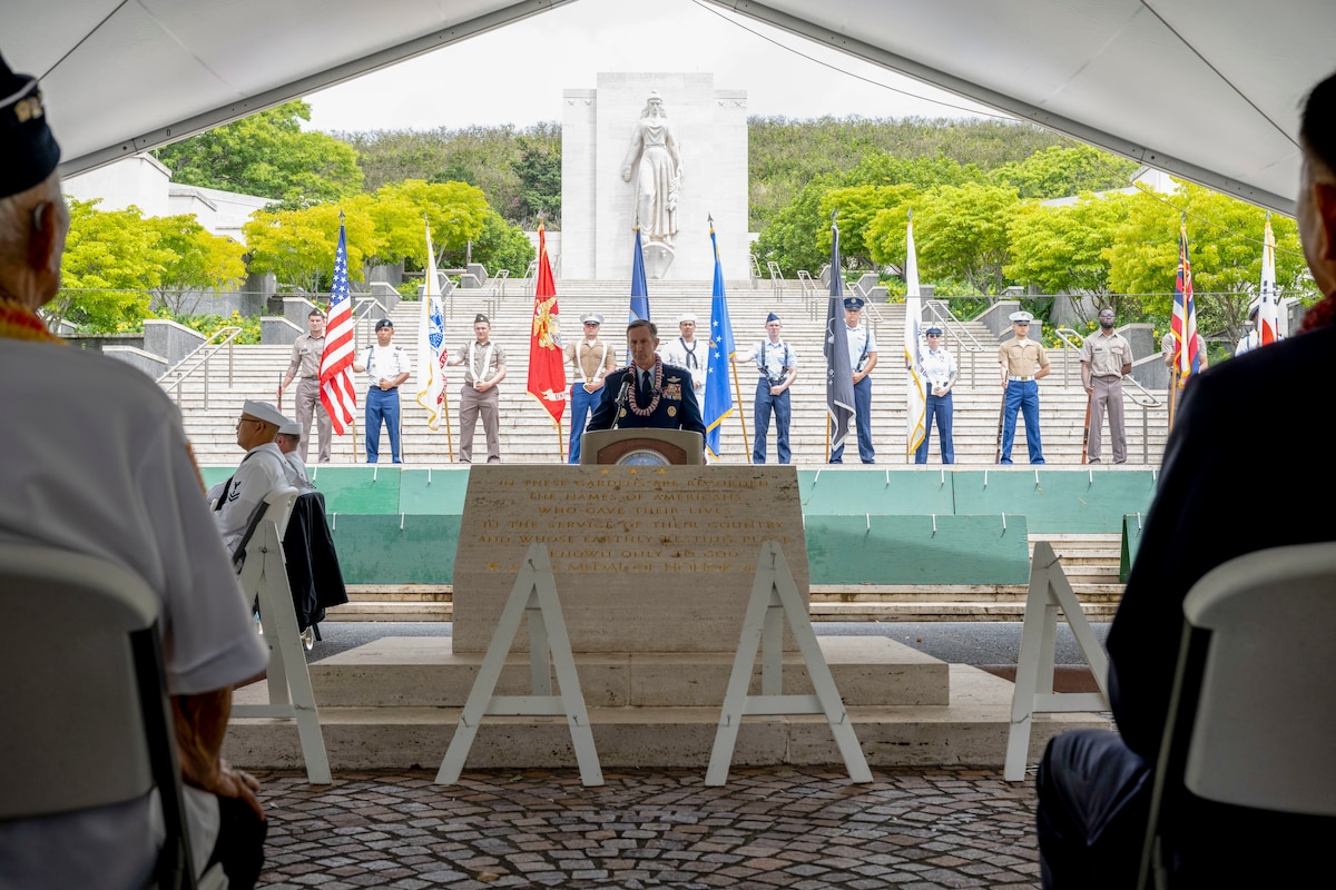 A uniformed man stands at a podium in front of a large crowd, with 12 soldiers lined up behind him holding flags.