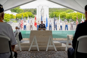 A uniformed man stands at a podium in front of a large crowd, with 12 soldiers lined up behind him holding flags.