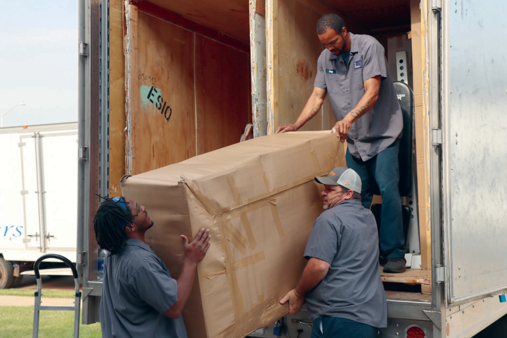 Three men dressed in casual attire unload a large item wrapped in brown paper off of a truck.