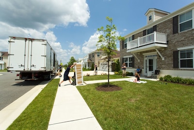 A man rolls a large item into a house while three people sit on the lawn and watch.
