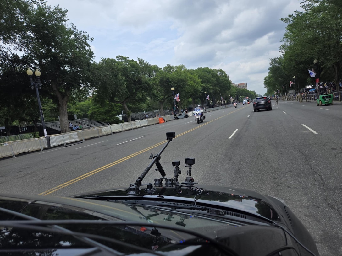 The view of a  hood of a car with multiple cameras and antennas as it goes down the middle of a street.