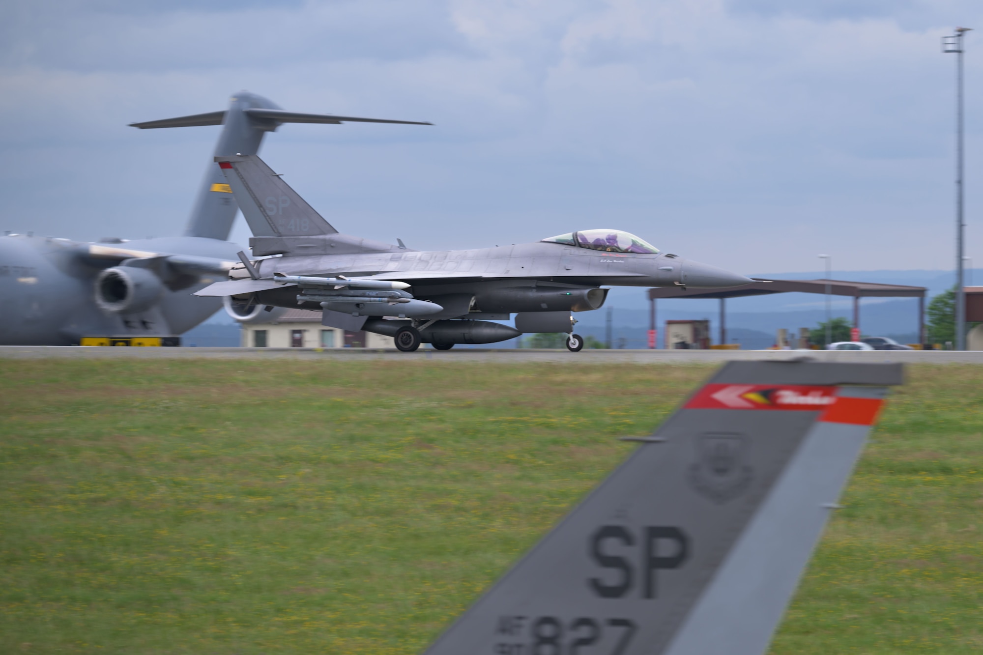 An F-16 fighter aircraft lands on the runway, passing two other aircraft on the flightline.
