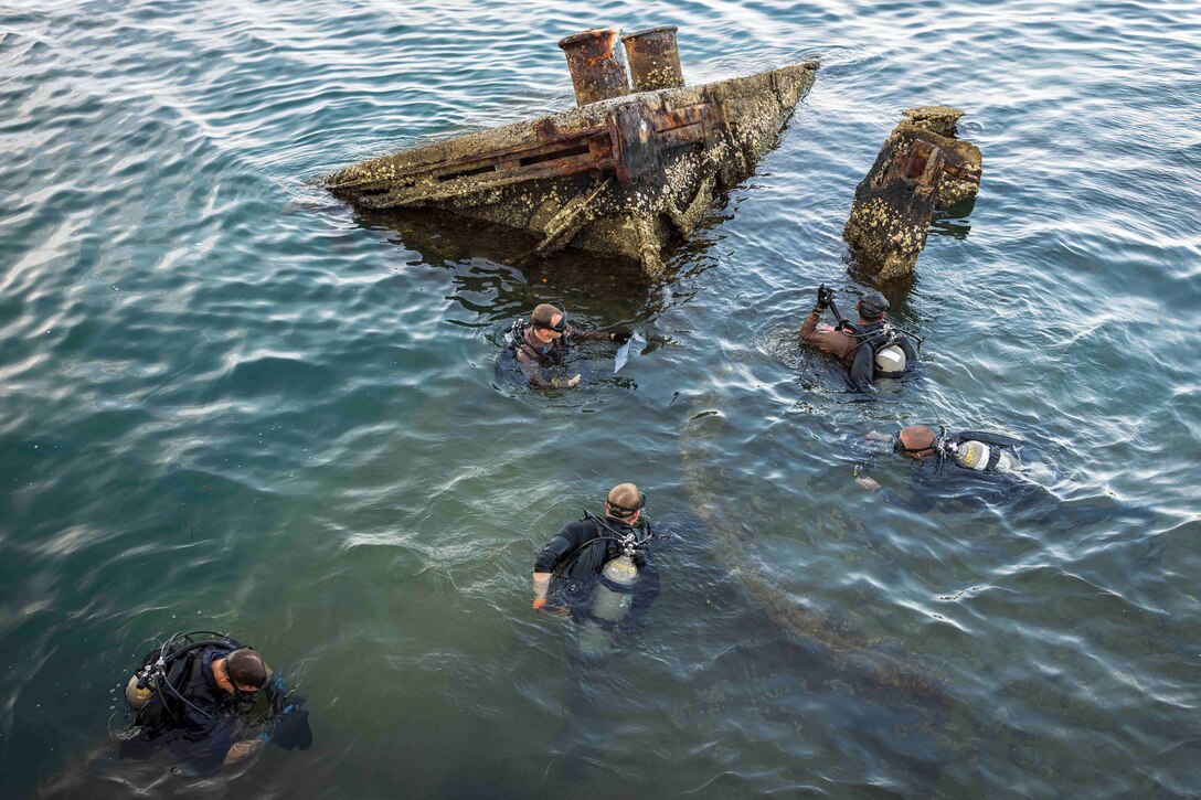 Divers in scuba gear swim around the wreckage of a ship submerged in murky water.