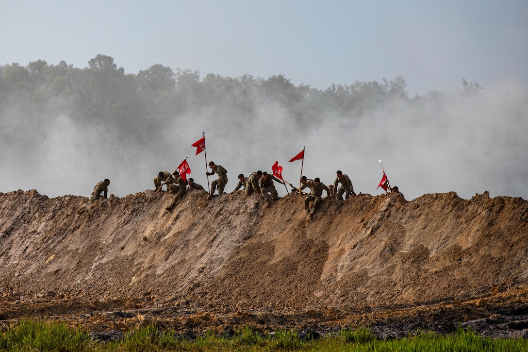 Soldiers carrying red flags climb over a dirt hill as smoke rises in the background.