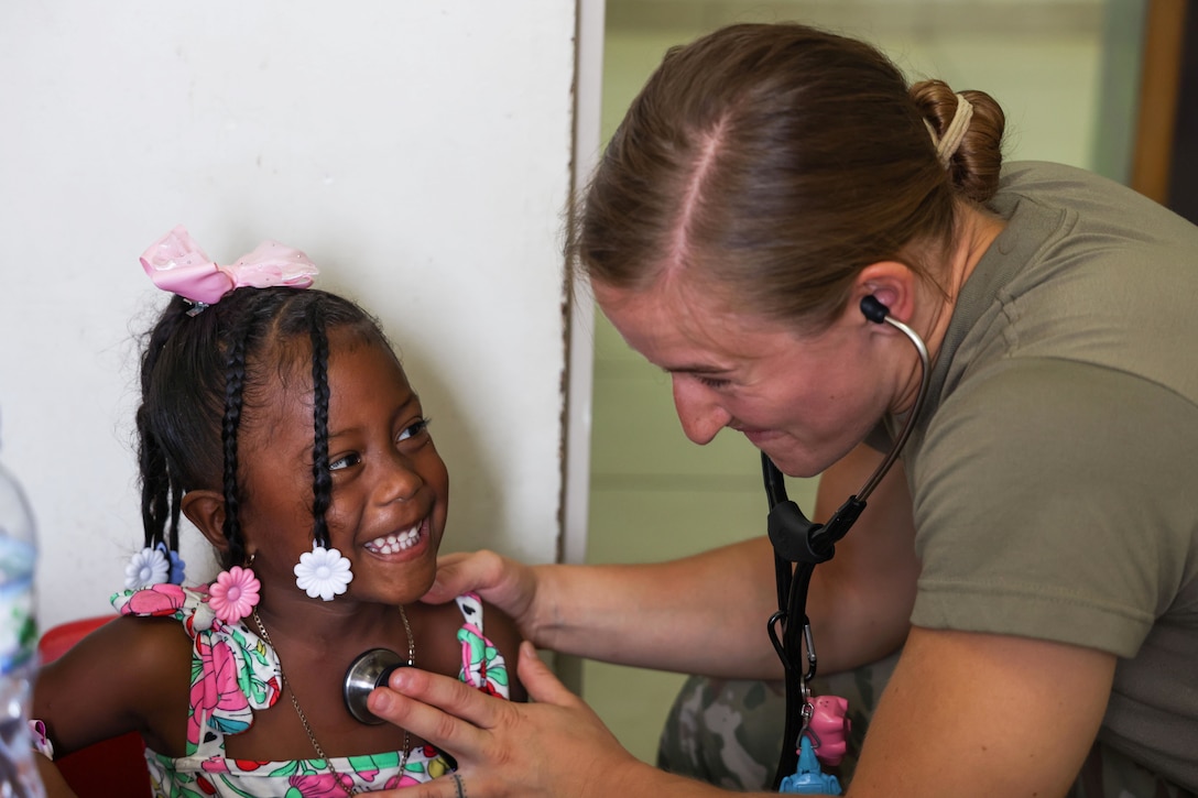 A child smiles at an airman as they place a stethoscope on their heart.