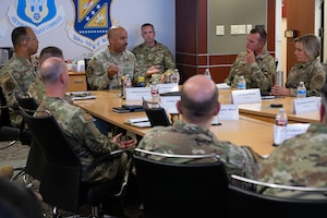 An individual in military uniform seated at the head of a table speaks to a seated group.