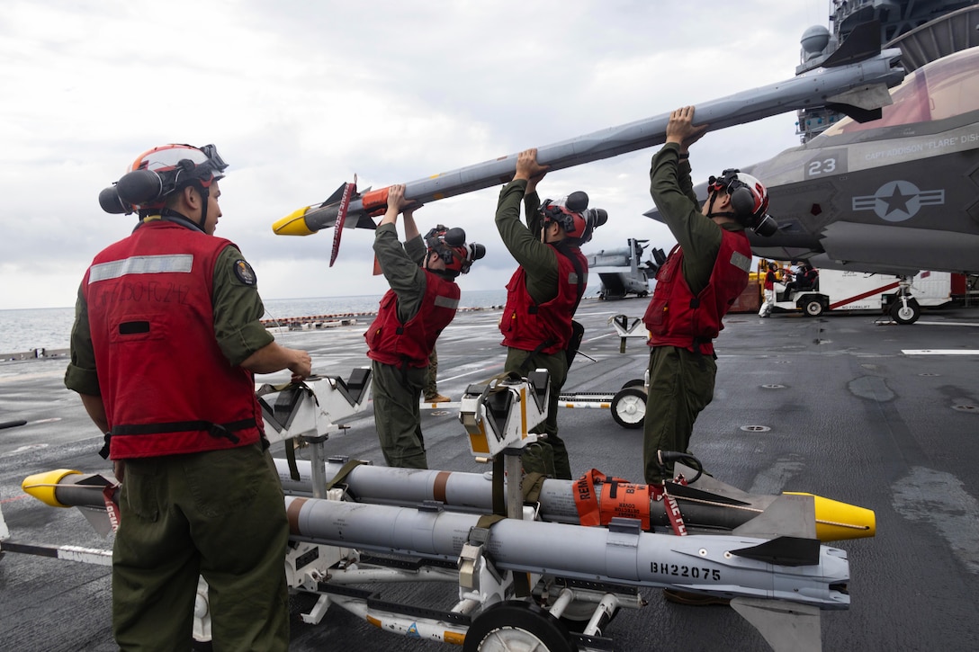 Three Marines stand near a partially visible aircraft aboard a ship as they carry ammo while a fellow Marine watches.