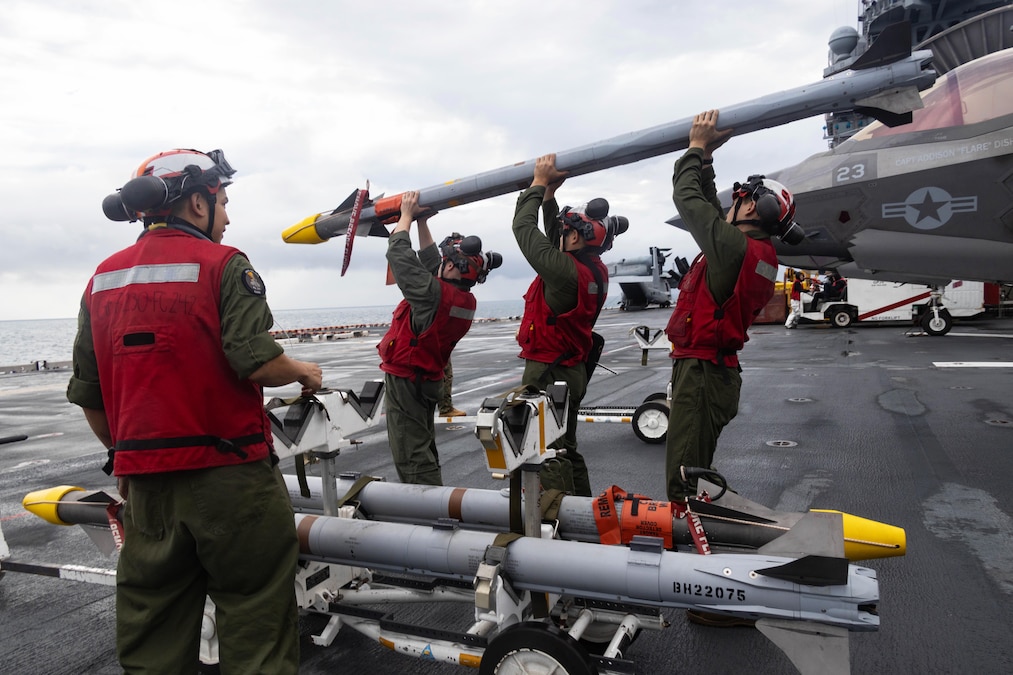 Three Marines stand near a partially visible aircraft aboard a ship as they carry ammo while a fellow Marine watches.