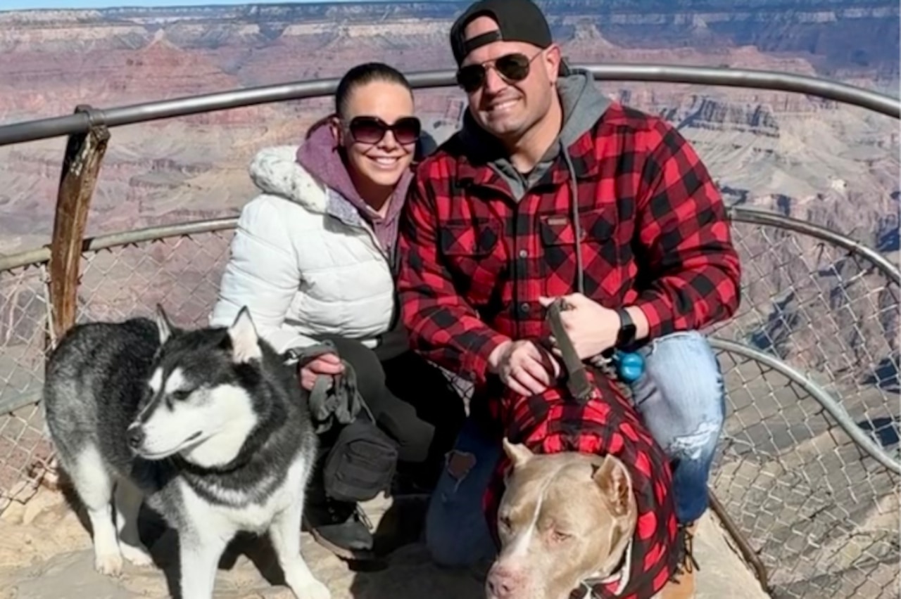 Two people pose with two dogs on an overlook overlooking a canyon.
