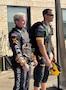Two men in skydiving apparel look off in the distance at a plane (not shown)