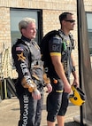 Two men in skydiving apparel look off in the distance at a plane (not shown)