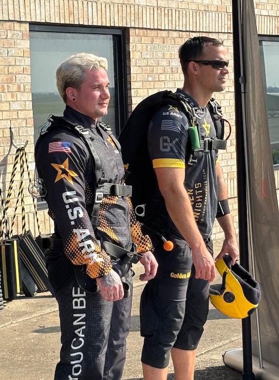 Two men in skydiving apparel look off in the distance at a plane (not shown)