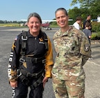 A woman in a skydiving flight suit stands posed and smiling next to a female in an Army outfit outside near a small airport entrance