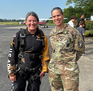 A woman in a skydiving flight suit stands posed and smiling next to a female in an Army outfit outside near a small airport entrance