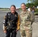 A woman in a skydiving flight suit stands posed and smiling next to a female in an Army outfit outside near a small airport entrance