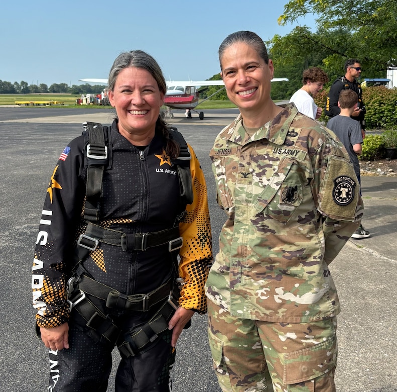 A woman in a skydiving flight suit stands posed and smiling next to a female in an Army outfit outside near a small airport entrance