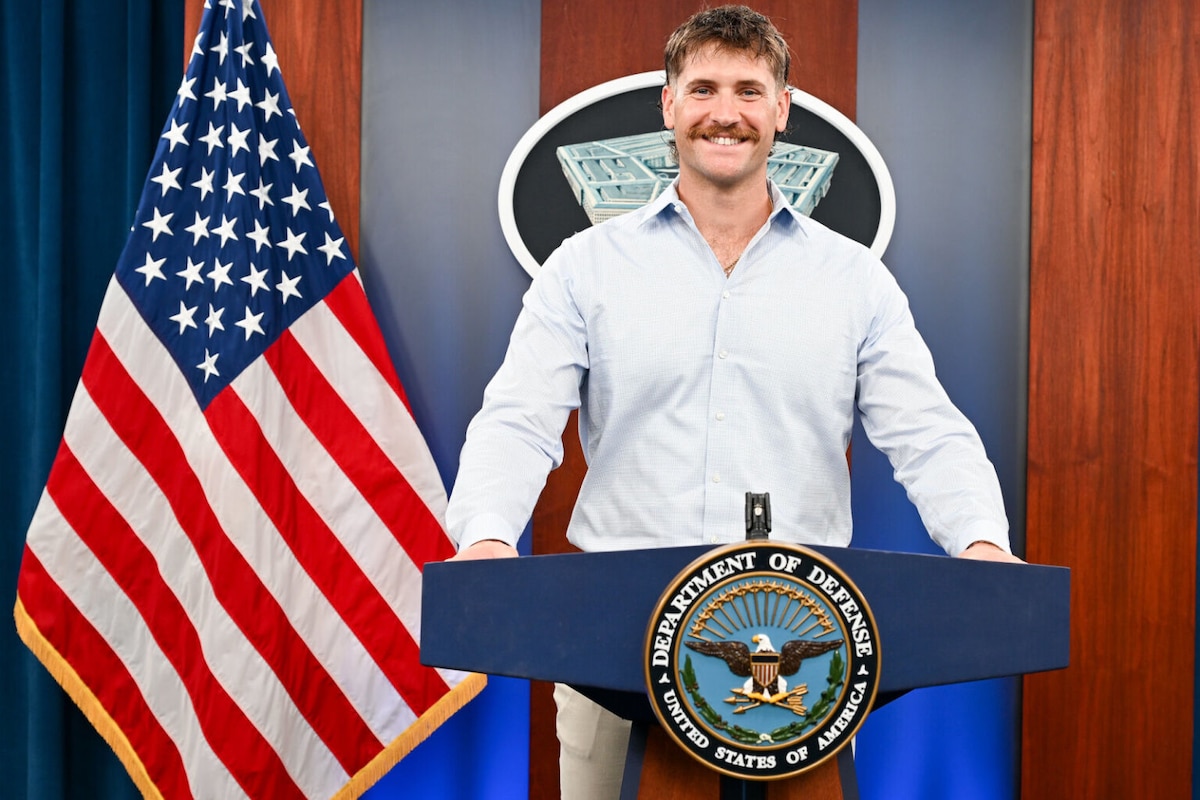 A man in casual attire is standing behind a podium smiling with a logo that reads “Department of Defense – United States of America.” An American flag is on his left.