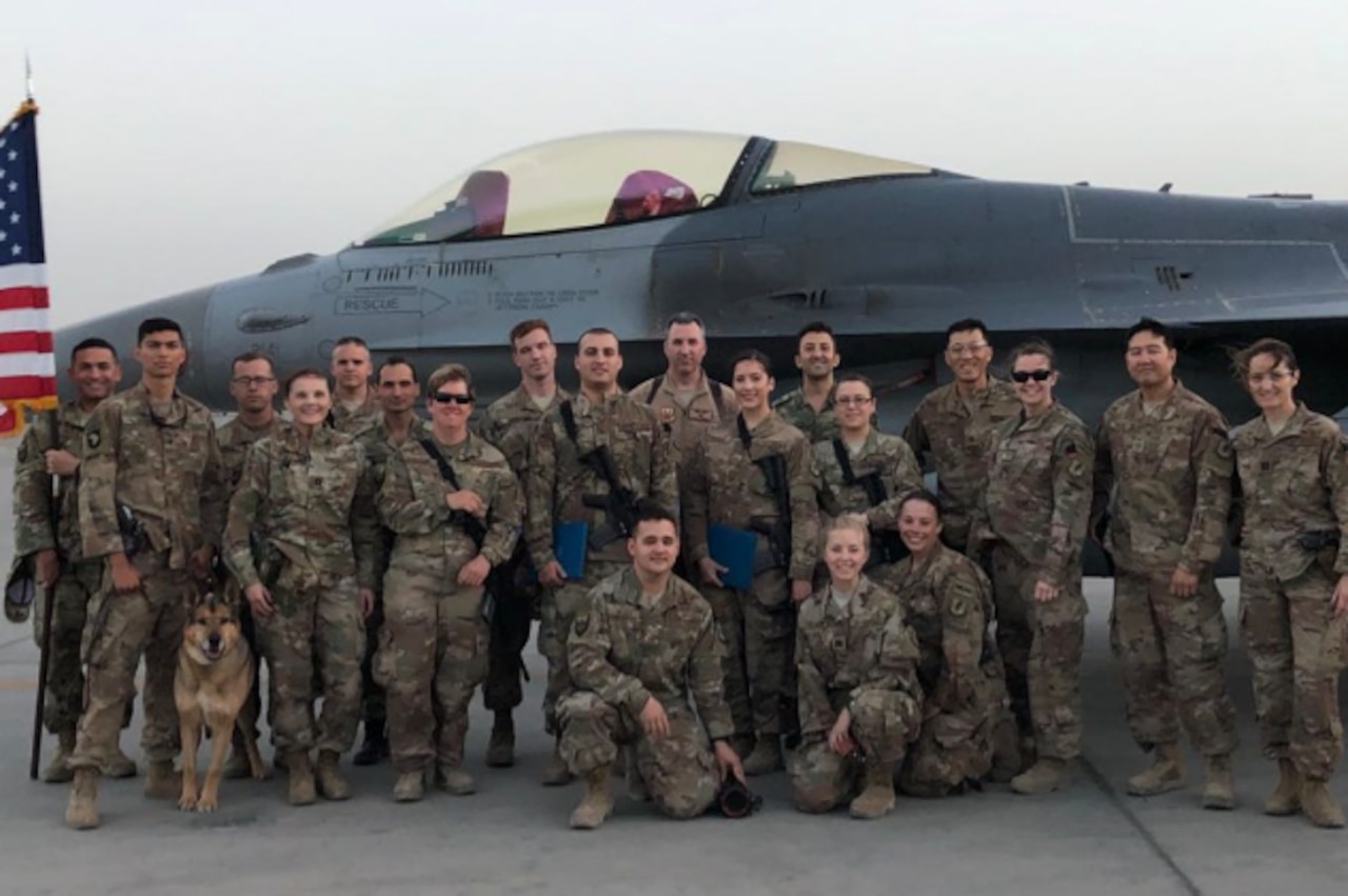 A group of service members and a dog pose for a photo in front of an aircraft on the flight line.