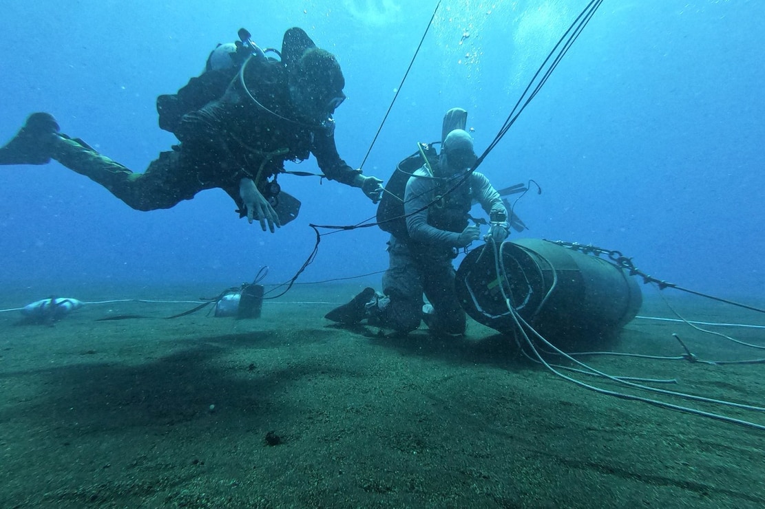 Two sailors in diving gear attach wires to a mine in deep blue waters.