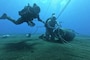 Two sailors in diving gear attach wires to a mine in deep blue waters.
