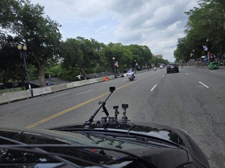 The view of a  hood of a car with multiple cameras and antennas as it goes down the middle of a street.