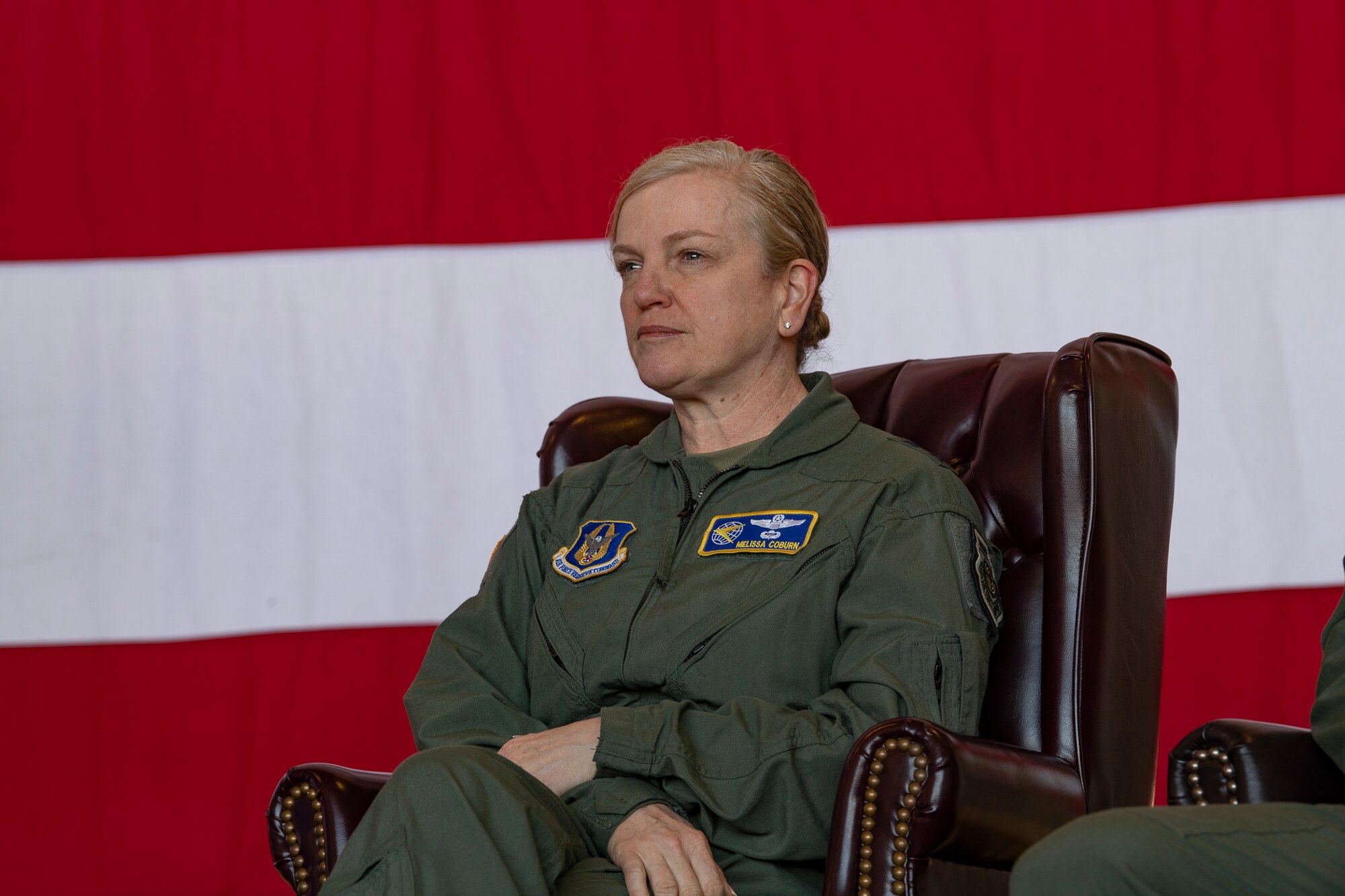 A woman in a flight suit sitting on stage in front of an American flag.