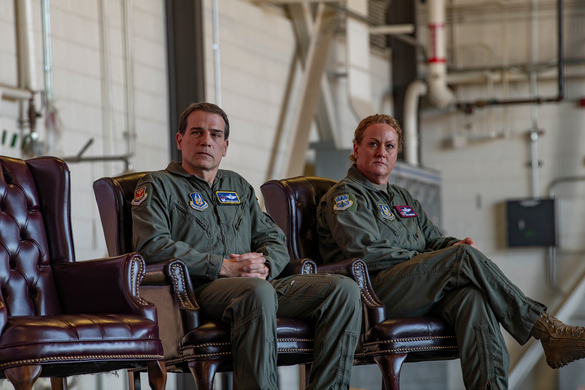 Two people in flight suits sitting on a chair on stage.