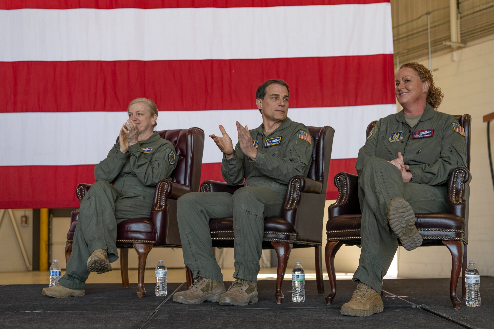 Three people in flight suits sit on a stage in front of an American flag.