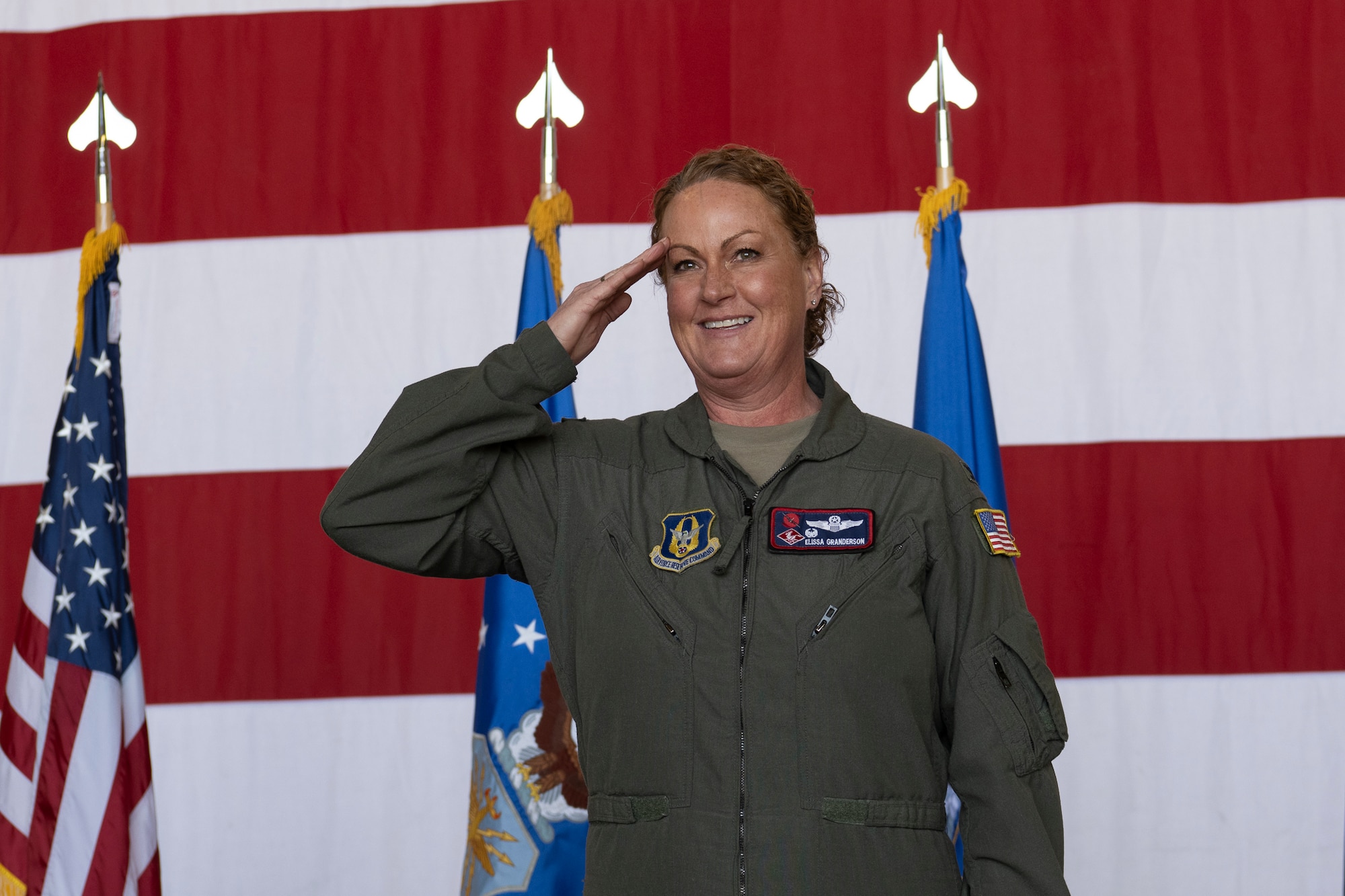 A woman in a military flight suit renders a salute with the American flag behind her.