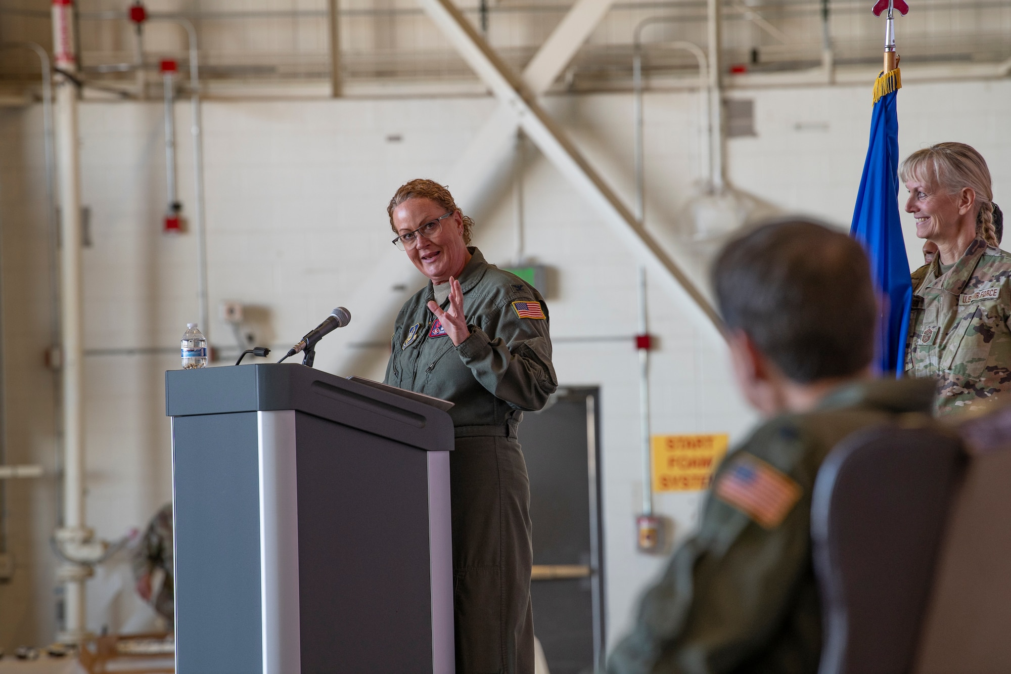 A woman in a flight suit on a stage speaks to a man in a flight suit sitting down.
