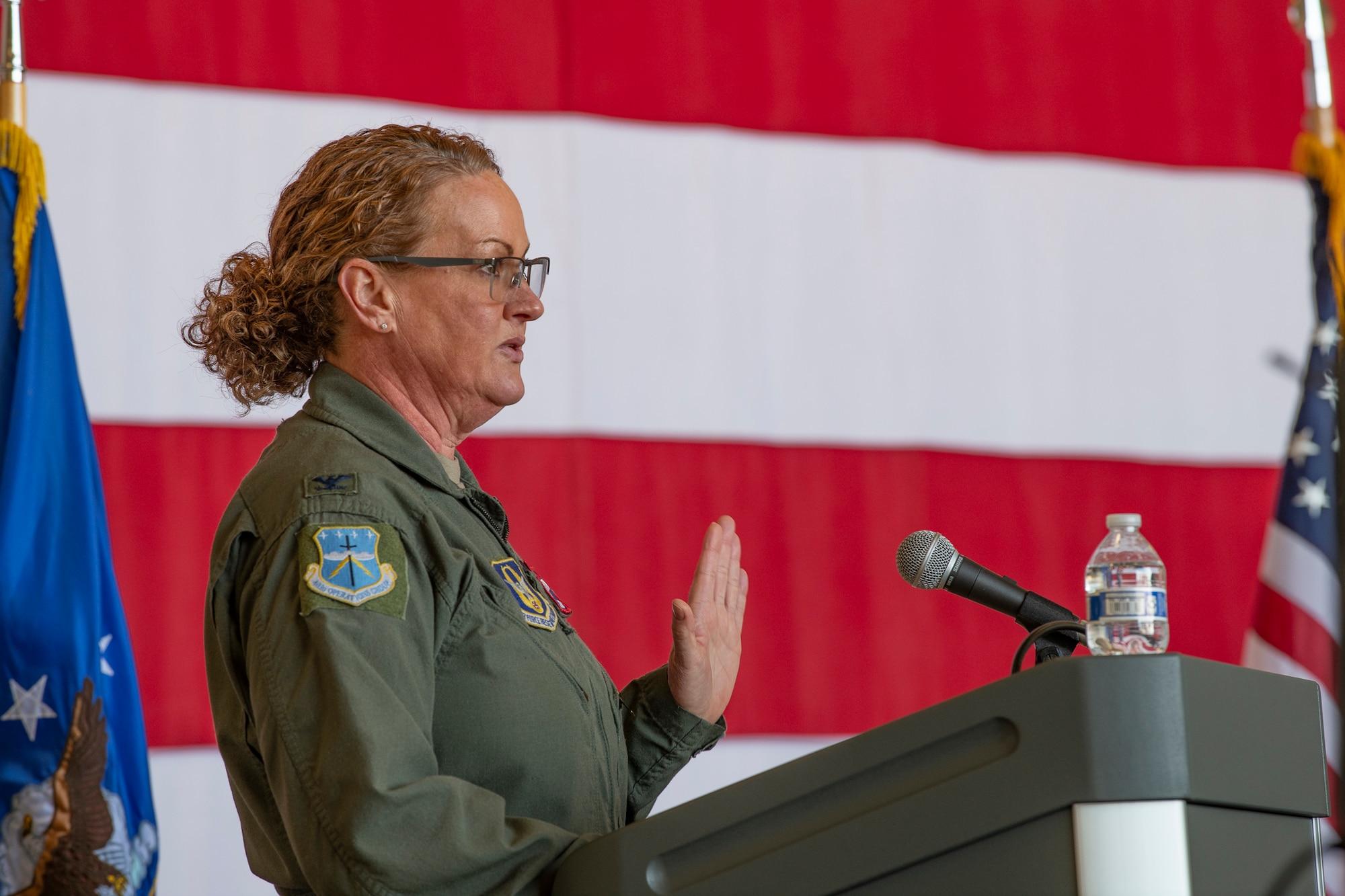 A woman in a flight suit stands in front of a podium on a stage  with an American flag in the background.