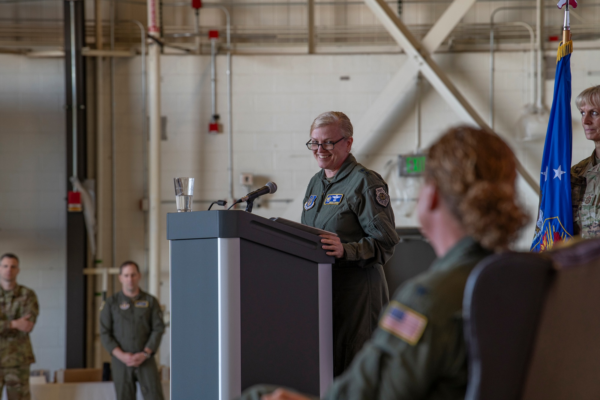 Two women in flight suits on stage, one at a podium speaking to the other.