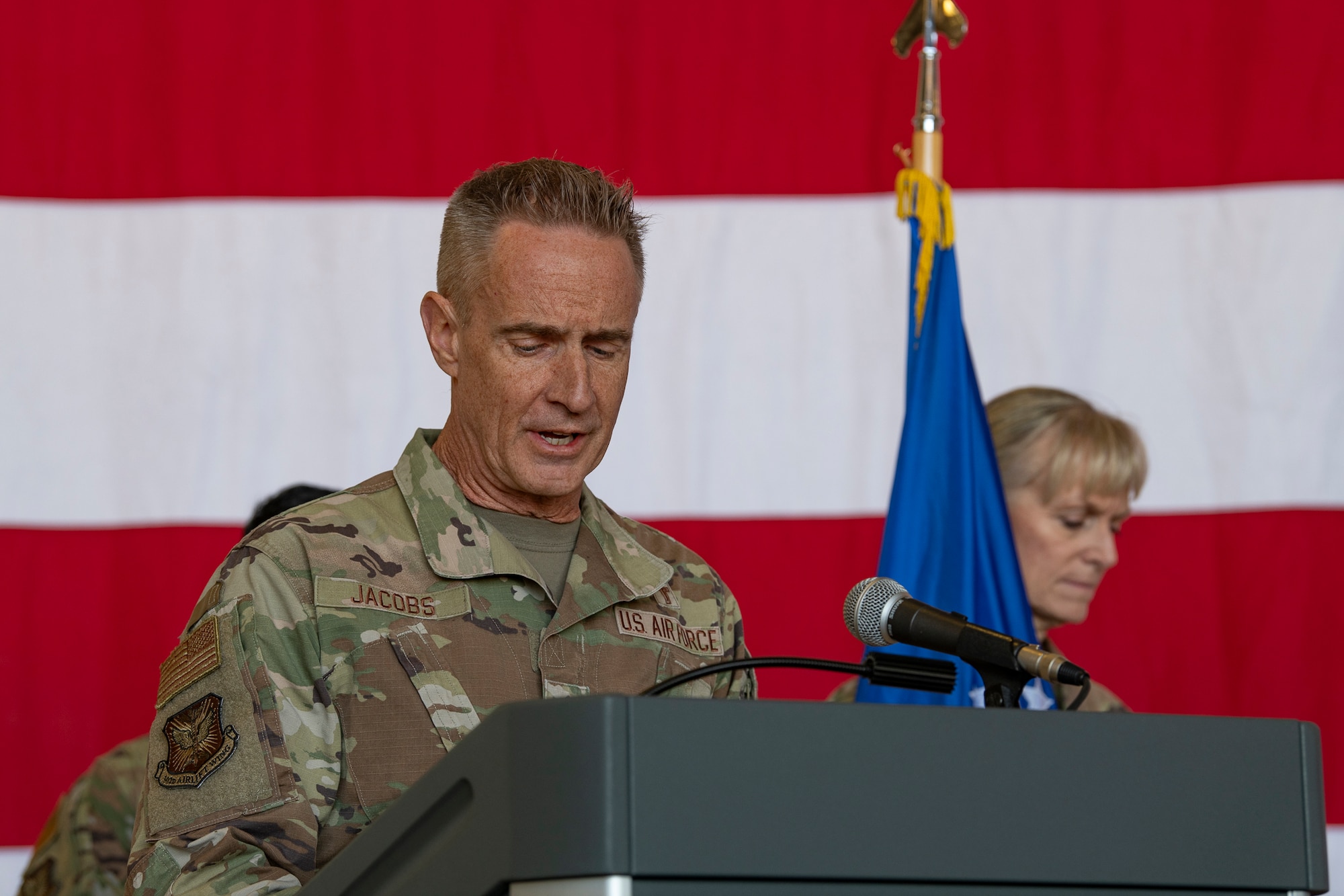 A man in military uniform speaks on stage with an American flag in the background.