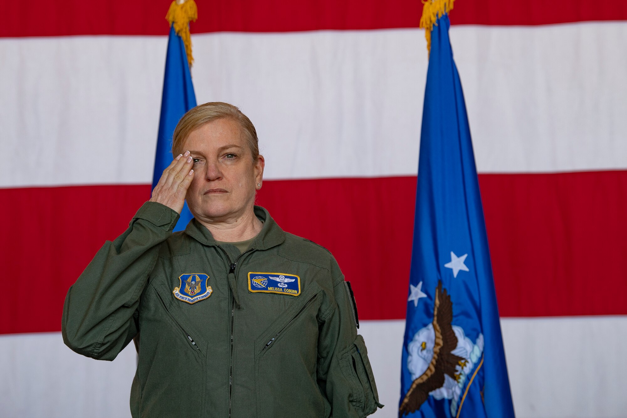 A woman in a flight suit salutes on stage with an American flag behind her.