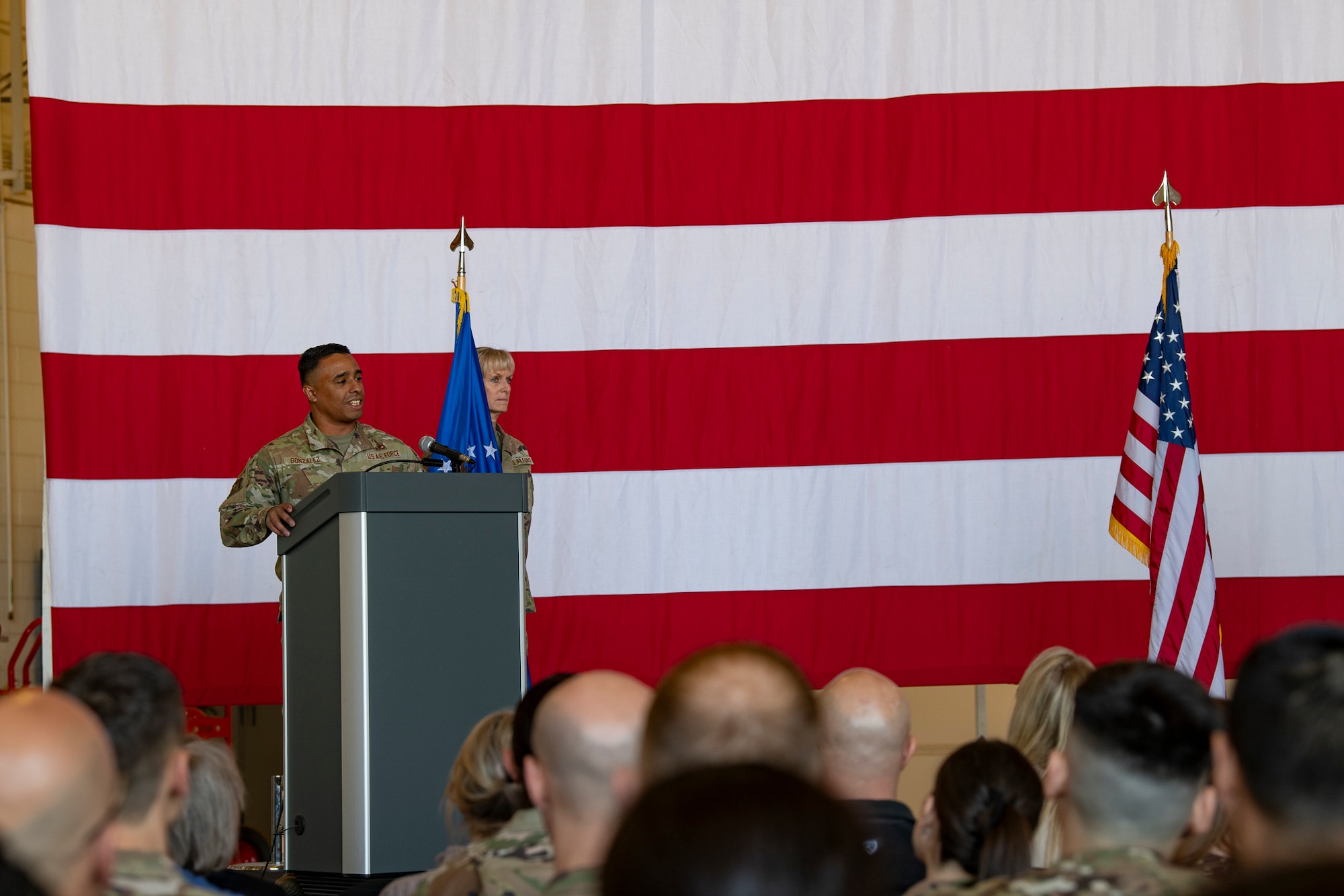 A man on stage speaks to a crowd with an American flag in the background.