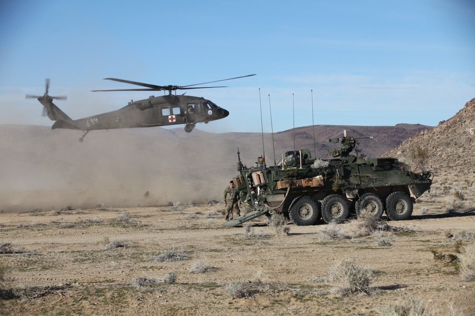 A helicopter hovers near a wheeled tactical vehicle in the high desert.