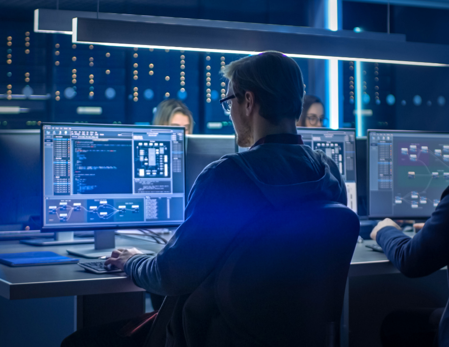A person in a dimly lit control room works on multiple computer monitors displaying complex data.