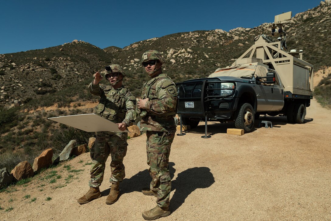 Soldiers stand on a dirt road in front of a parked vehicle with a mountain in the background.