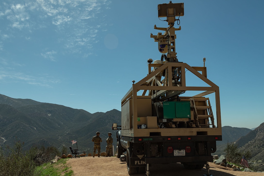 Soldiers stand on the dirt road near a large piece of construction equipment with mountains in the distance under a blue sky.